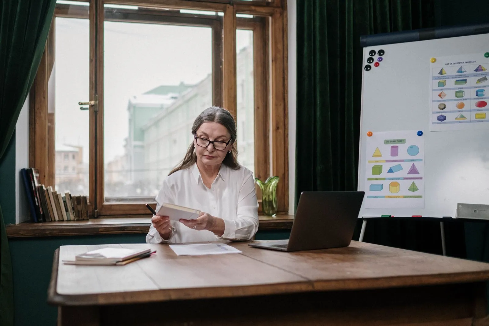 Elderly woman with glasses reviewing papers at a desk with laptop and charts.